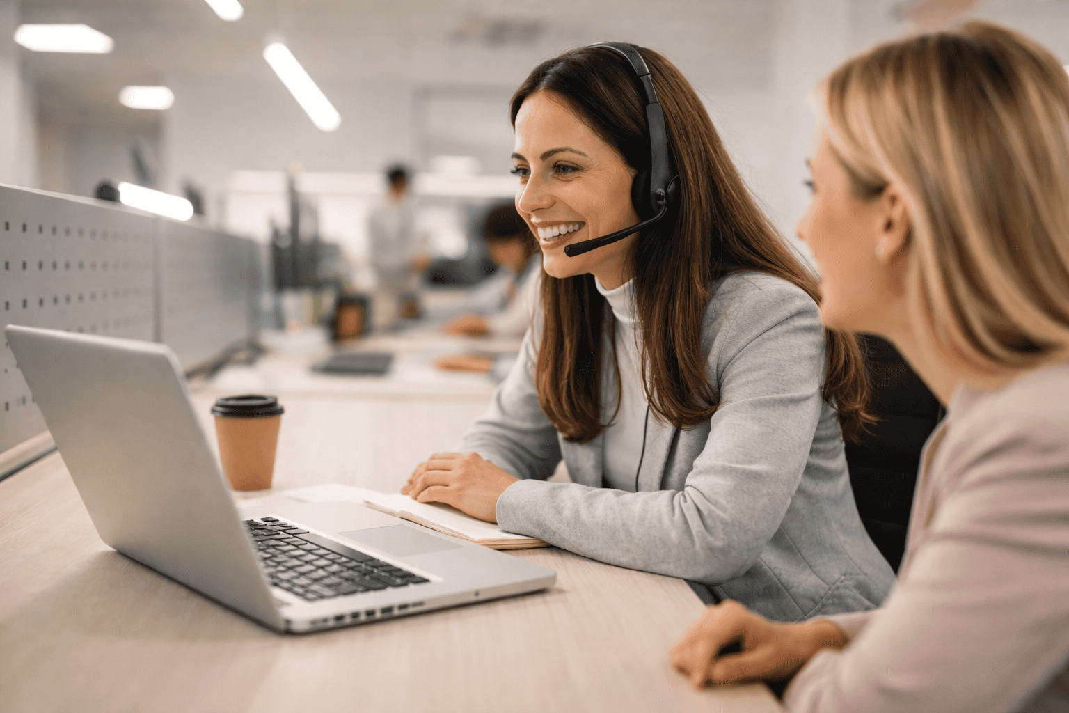 Conseillère en service client avec casque devant un ordinateur portable, échangeant avec une collègue dans un bureau moderne