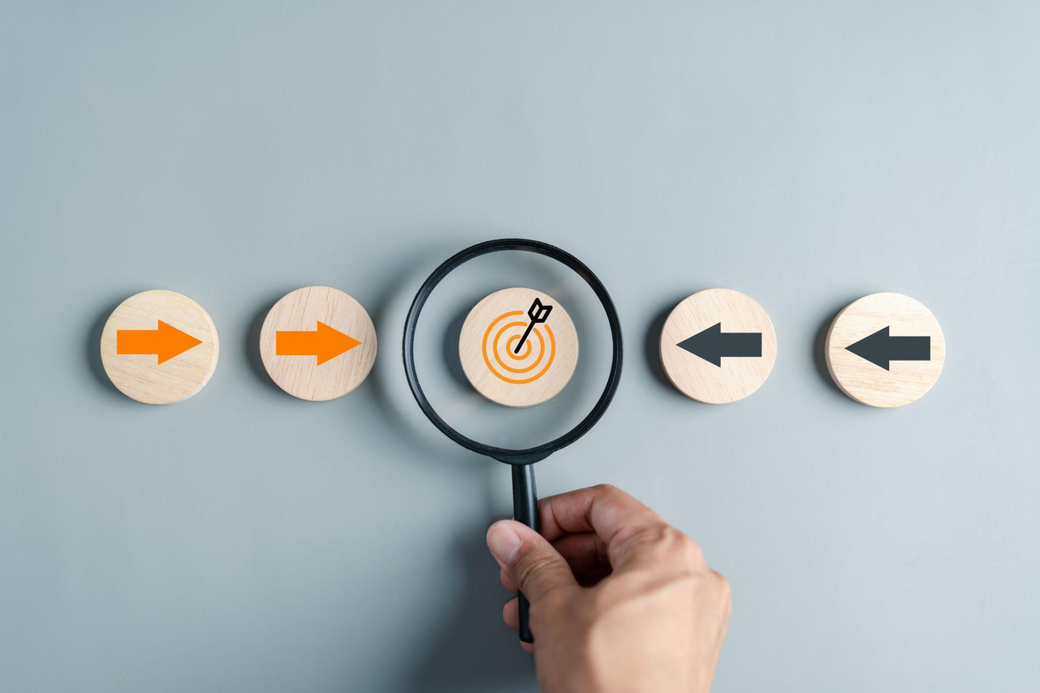 Main tenant une loupe centrée sur une cible parmi des flèches opposées, illustrant le positionnement stratégique et la différenciation sur le marché