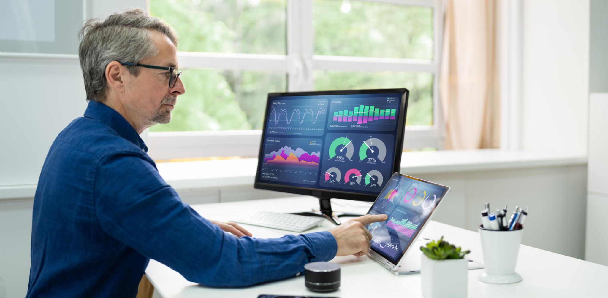 Homme analysant des tableaux de bord sur un écran et une tablette, affichant des graphiques et indicateurs de performance pour le suivi de l’activité